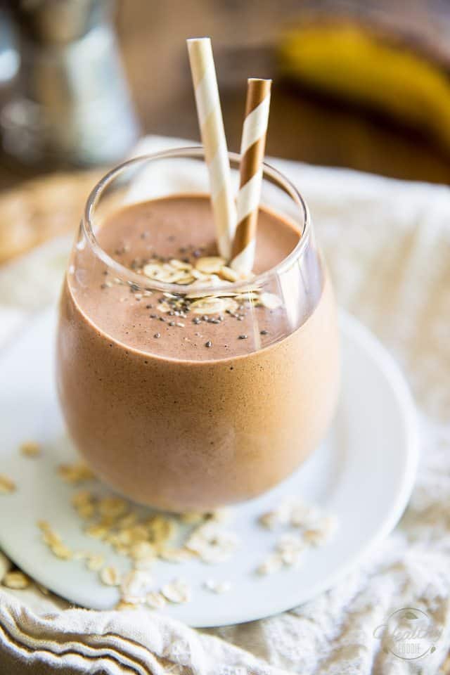 A glass of chocolate smoothie topped with oats and chia seeds, served with two striped straws on a white plate—perfect inspiration for those seeking high-protein smoothie recipes.