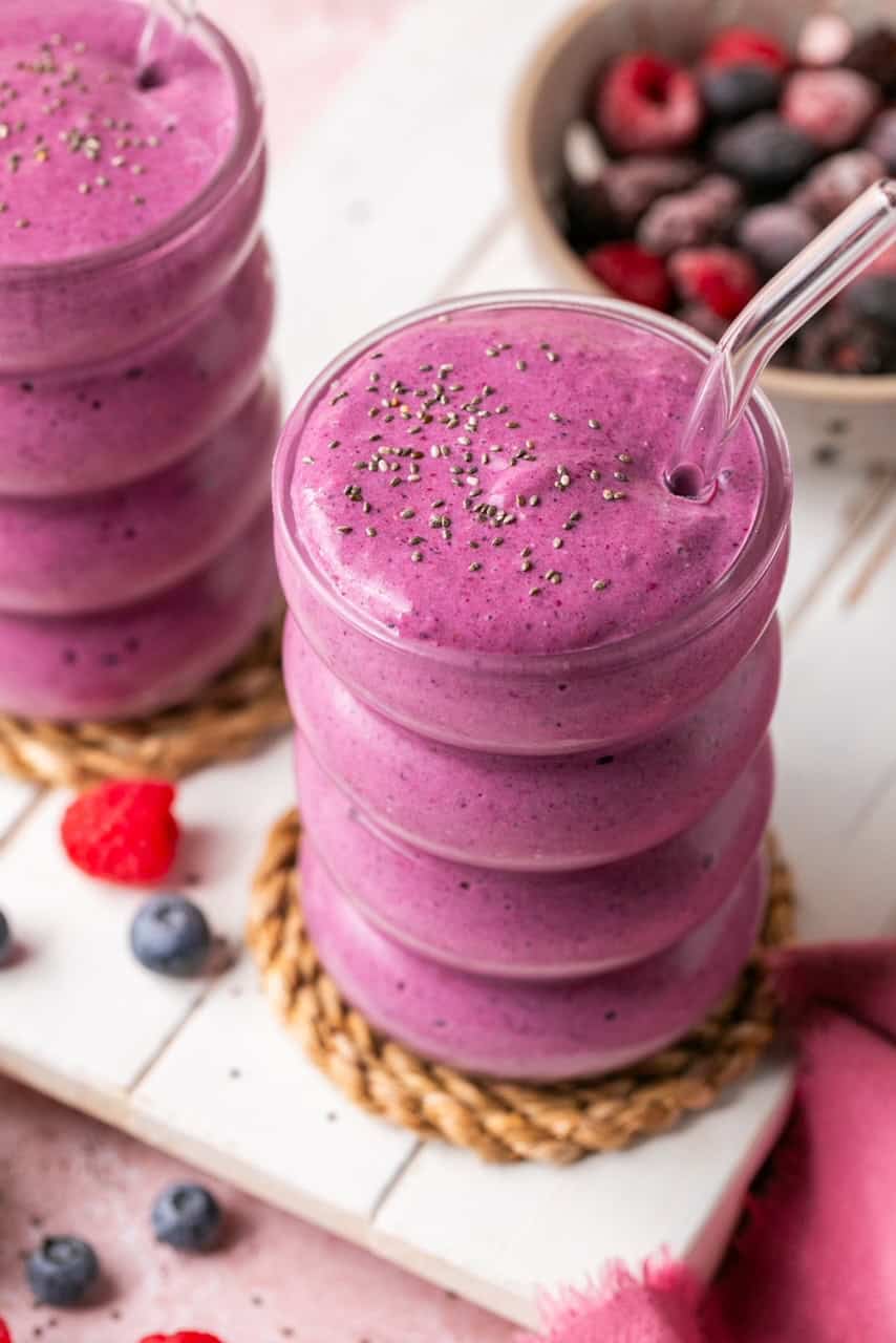 A glass of thick purple smoothie, inspired by delicious Smoothie Recipes, with chia seeds on top and a clear straw, is surrounded by scattered berries and a bowl of mixed berries in the background.