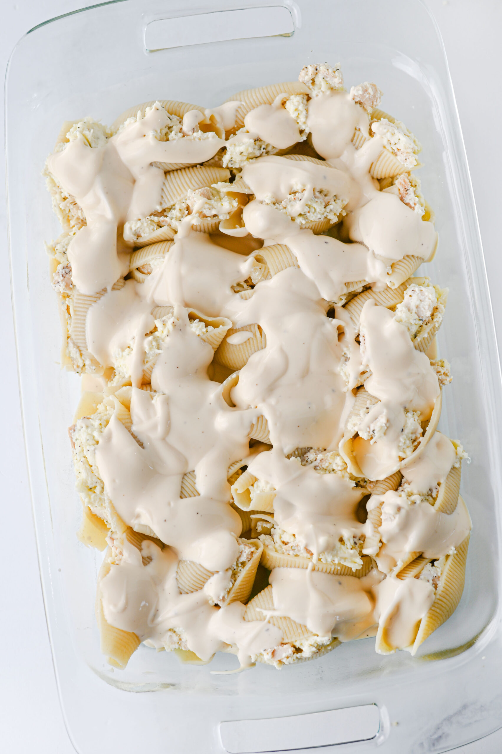 Alfredo sauce poured over chicken stuffed shells in a baking dish.