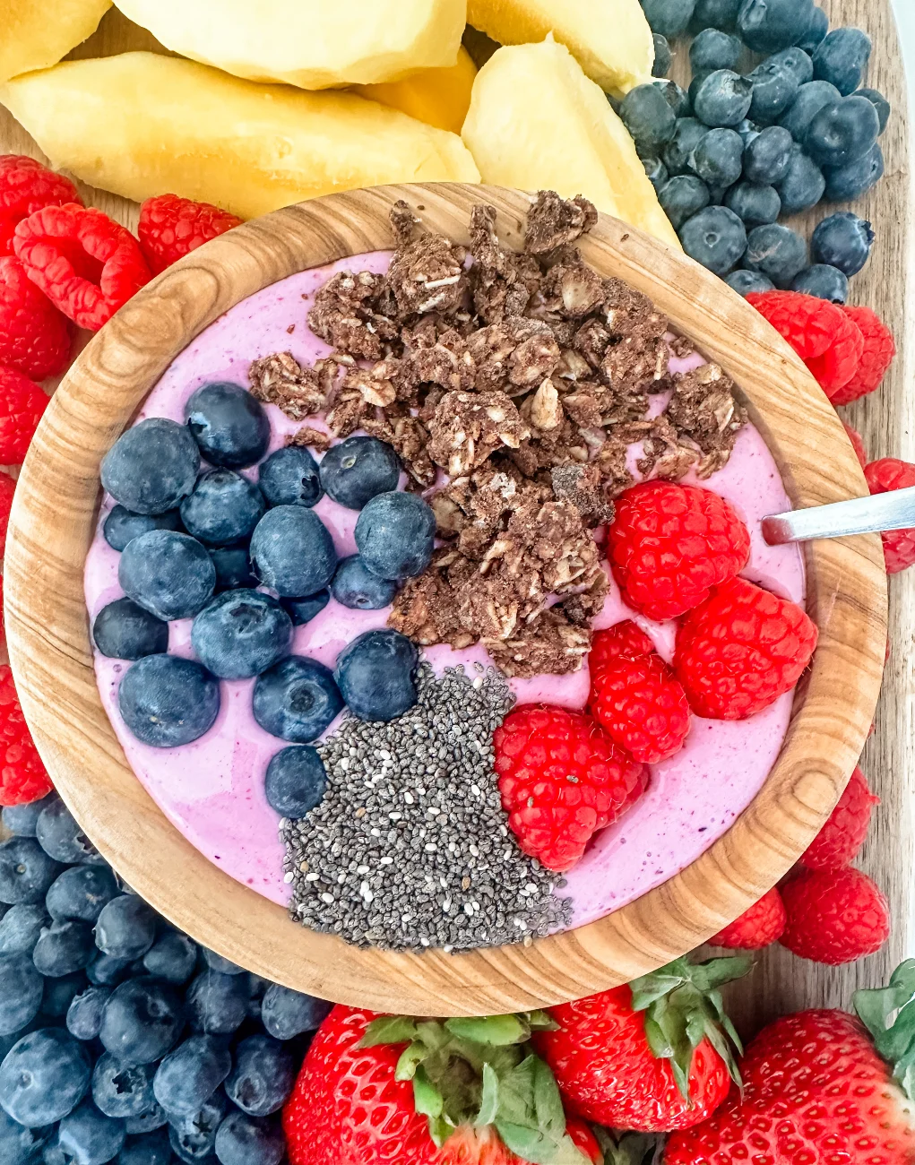 A smoothie bowl topped with granola, blueberries, raspberries, and chia seeds, surrounded by fresh strawberries, raspberries, blueberries, and pineapple slices—perfect inspiration for anyone seeking delicious protein smoothies or high-protein smoothie recipes.