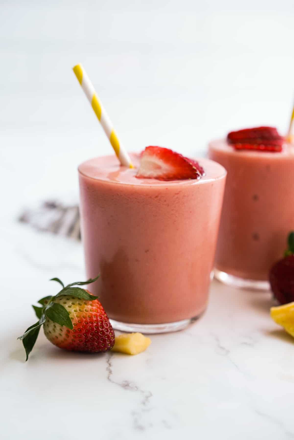 Two glasses of pink smoothie with yellow striped straws, garnished with sliced strawberries, sit on a white surface with a whole strawberry and a small pineapple piece nearby—an inviting scene perfect for anyone seeking delicious fruit smoothie recipes.