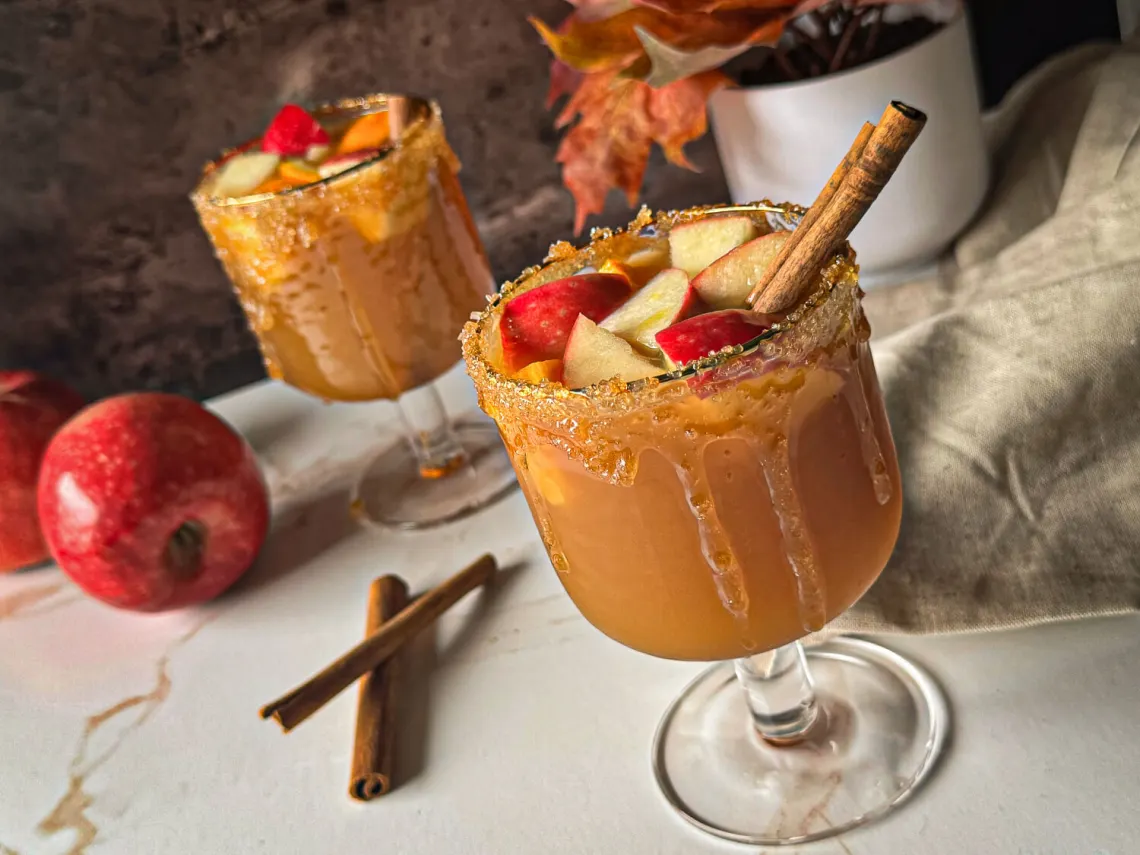 Two glasses of caramel apple cocktails garnished with apple slices and cinnamon sticks, with caramel-coated rims, sit on a white surface next to apples, cinnamon sticks, and autumn decor—perfect for cozy Autumn Gatherings.