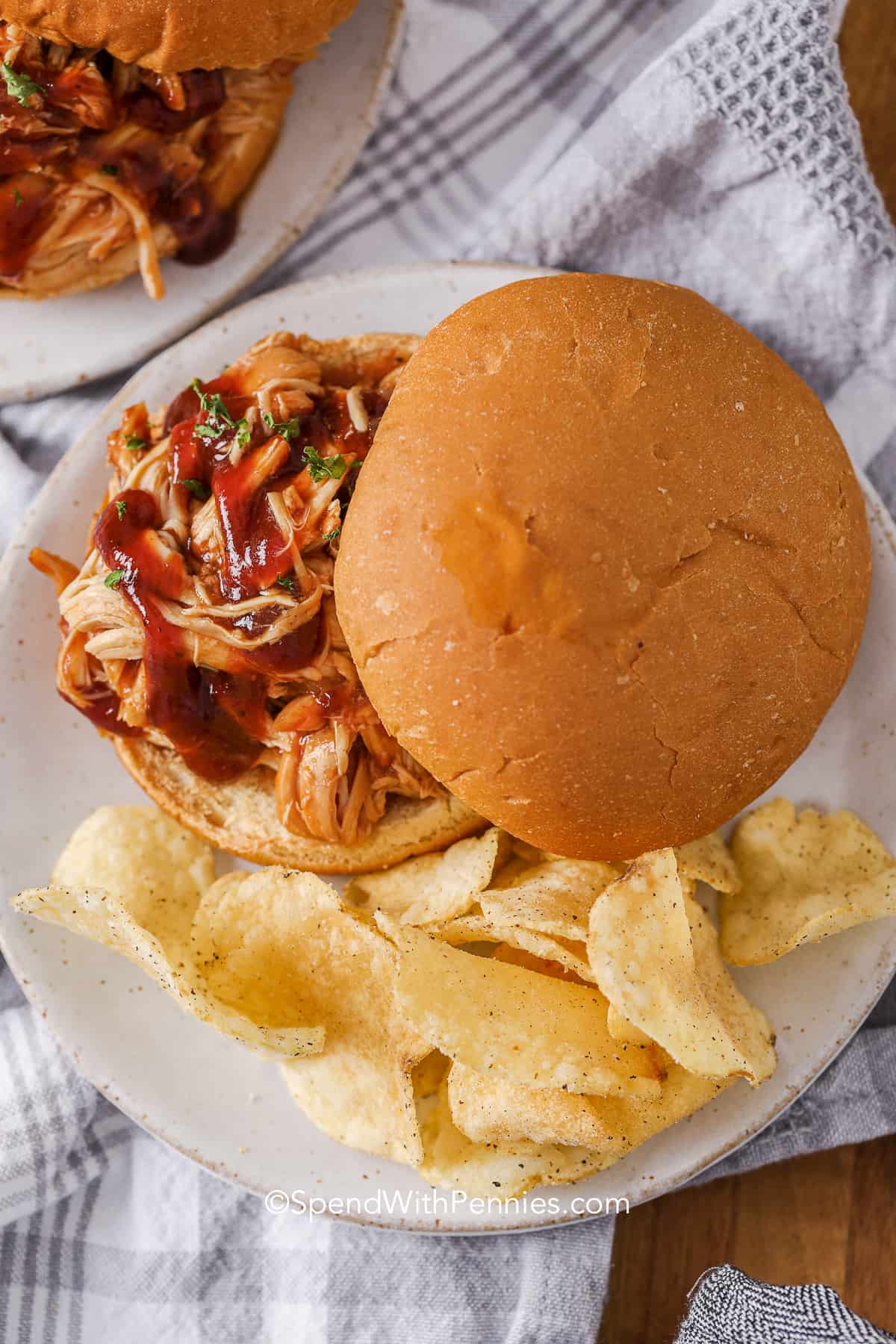 Barbecue pulled chicken sandwich on a bun served with potato chips on a white plate, placed on a striped cloth—perfect for easy dinners or to feed a crowd at team dinners.