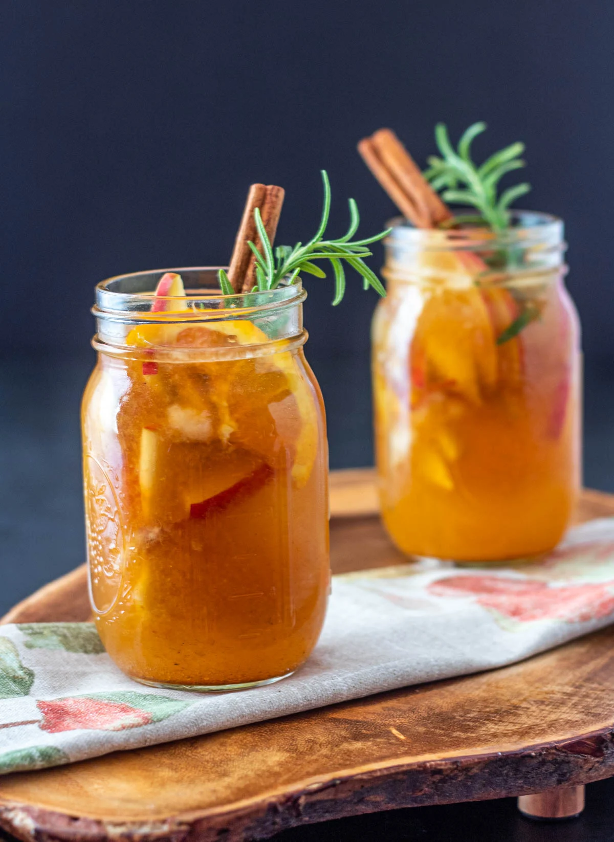Two mason jars filled with a cozy, orange-colored drink, garnished with apple slices, cinnamon sticks, and rosemary sprigs, placed on a wooden board with a floral napkin—perfect for autumn gatherings or as inspiration for your favorite fall sangria recipes.