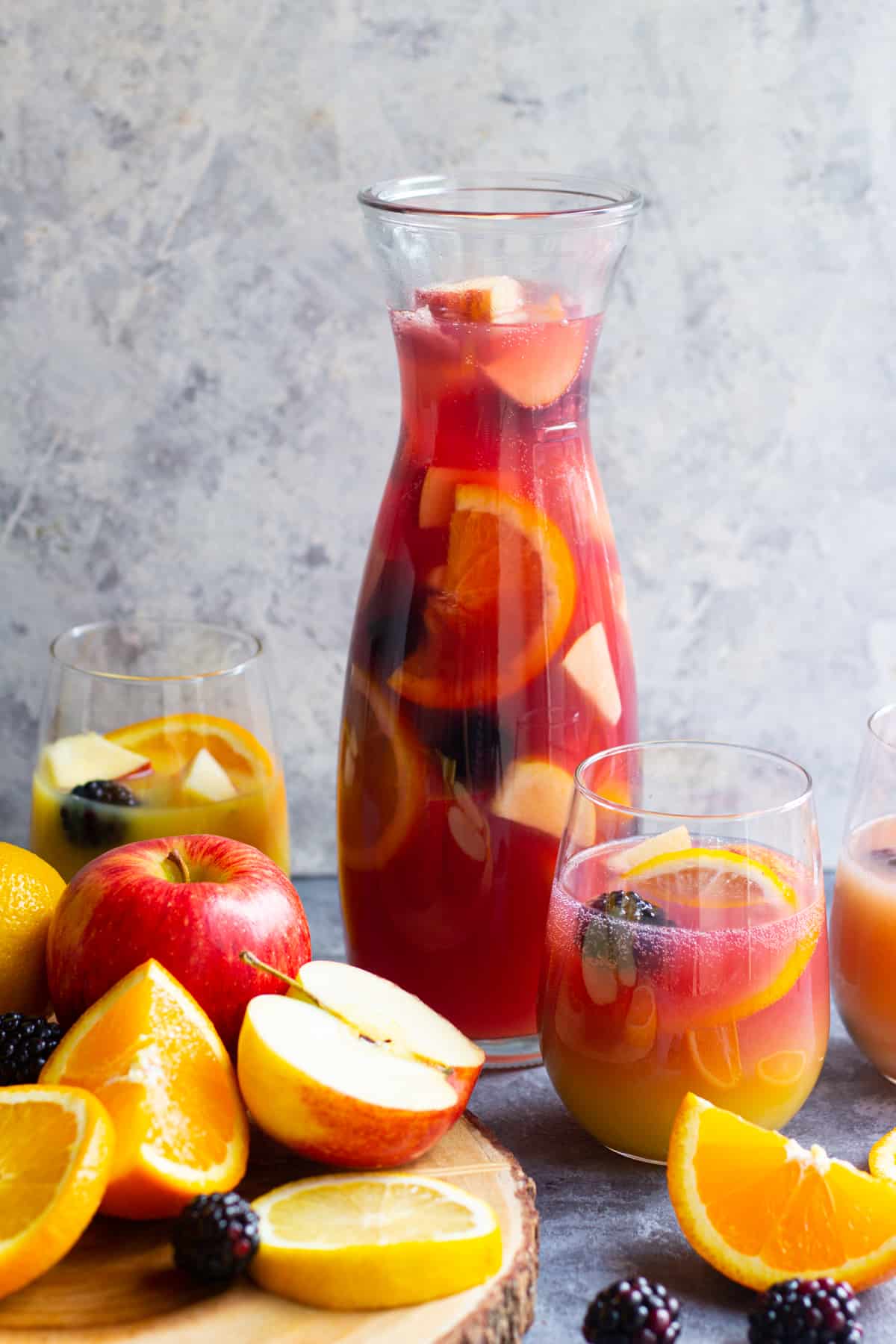 A glass pitcher and two glasses of fruit punch with sliced oranges, apples, and blackberries, surrounded by fresh fruit on a wooden board—perfect for Autumn Gatherings or as a cozy addition to your favorite Fall Sangria Recipes.