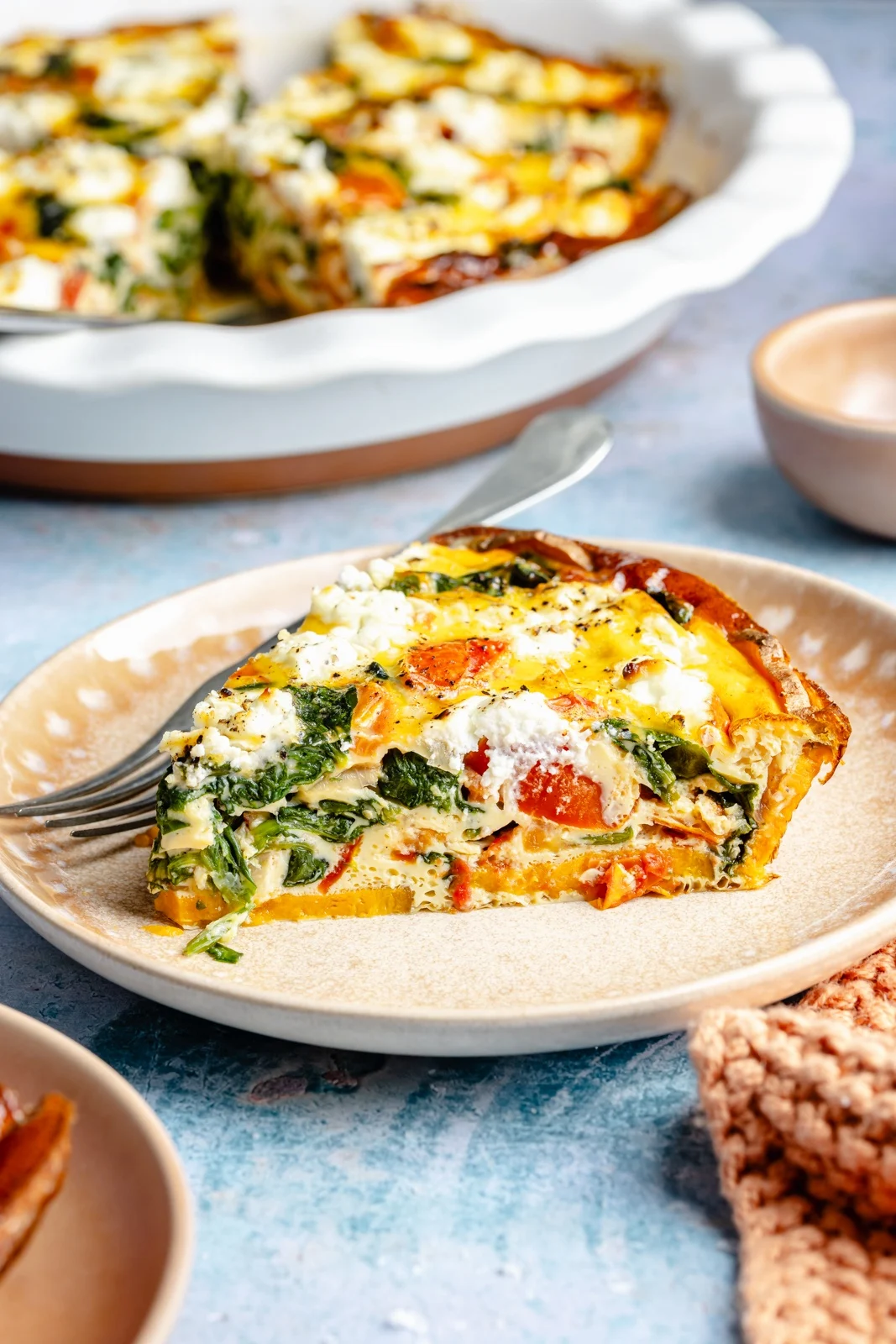 A slice of vegetable quiche with spinach, tomatoes, and cheese sits on a plate with a fork, with the remaining quiche in a pie dish in the background—perfect for Hoco brunch ideas or anyone looking to add delicious brunch food ideas to their menu.