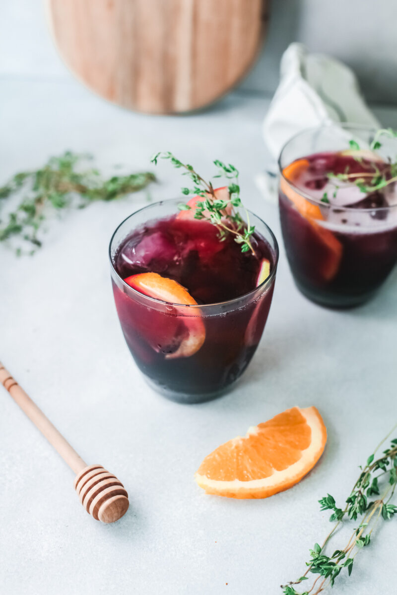 Two glasses of cozy dark red drink with ice, orange slices, and thyme sprigs sit invitingly next to an orange wedge, a honey dipper, and scattered herbs on a light surface—perfect for autumn gatherings or inspiring new fall sangria recipes.