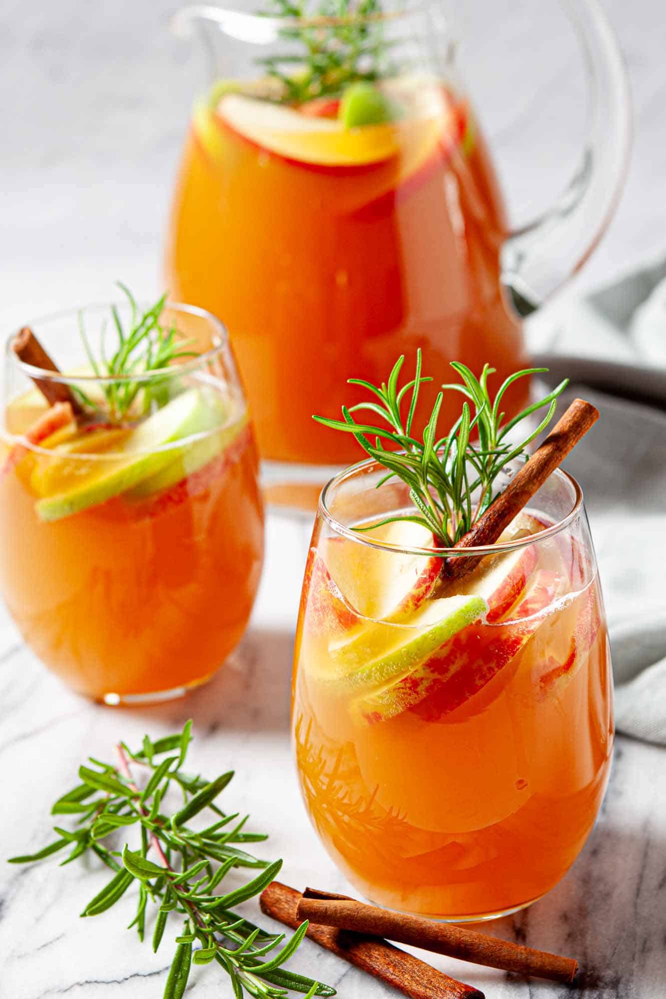 Two glasses and a pitcher of orange punch, garnished with apple slices, lime slices, rosemary sprigs, and cinnamon sticks on a marble surface—a cozy treat perfect for autumn gatherings or as inspiration for your favorite fall sangria recipes.
