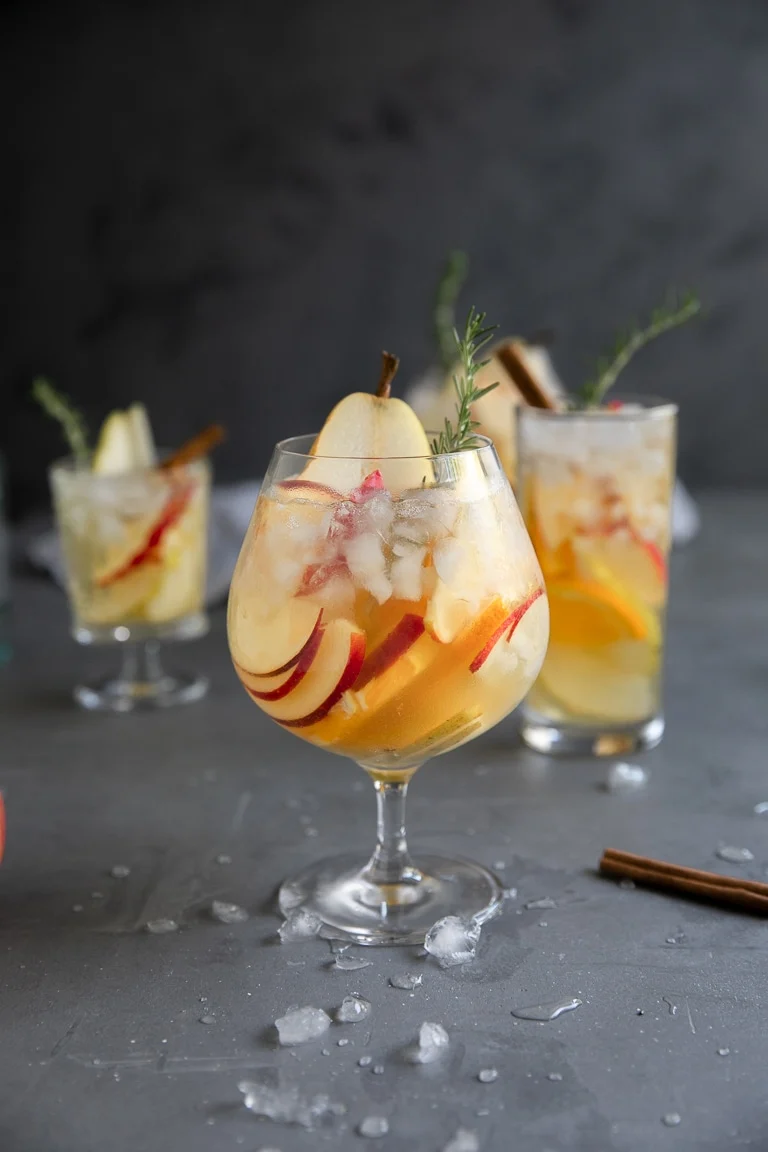 Three glasses of iced cocktail garnished with apple slices, pear slices, and rosemary sprigs are arranged on a gray surface with scattered ice cubes and a cinnamon stick—a perfect centerpiece for Autumn Gatherings or anyone seeking inspiration for cozy drinks and creative Fall Sangria recipes.