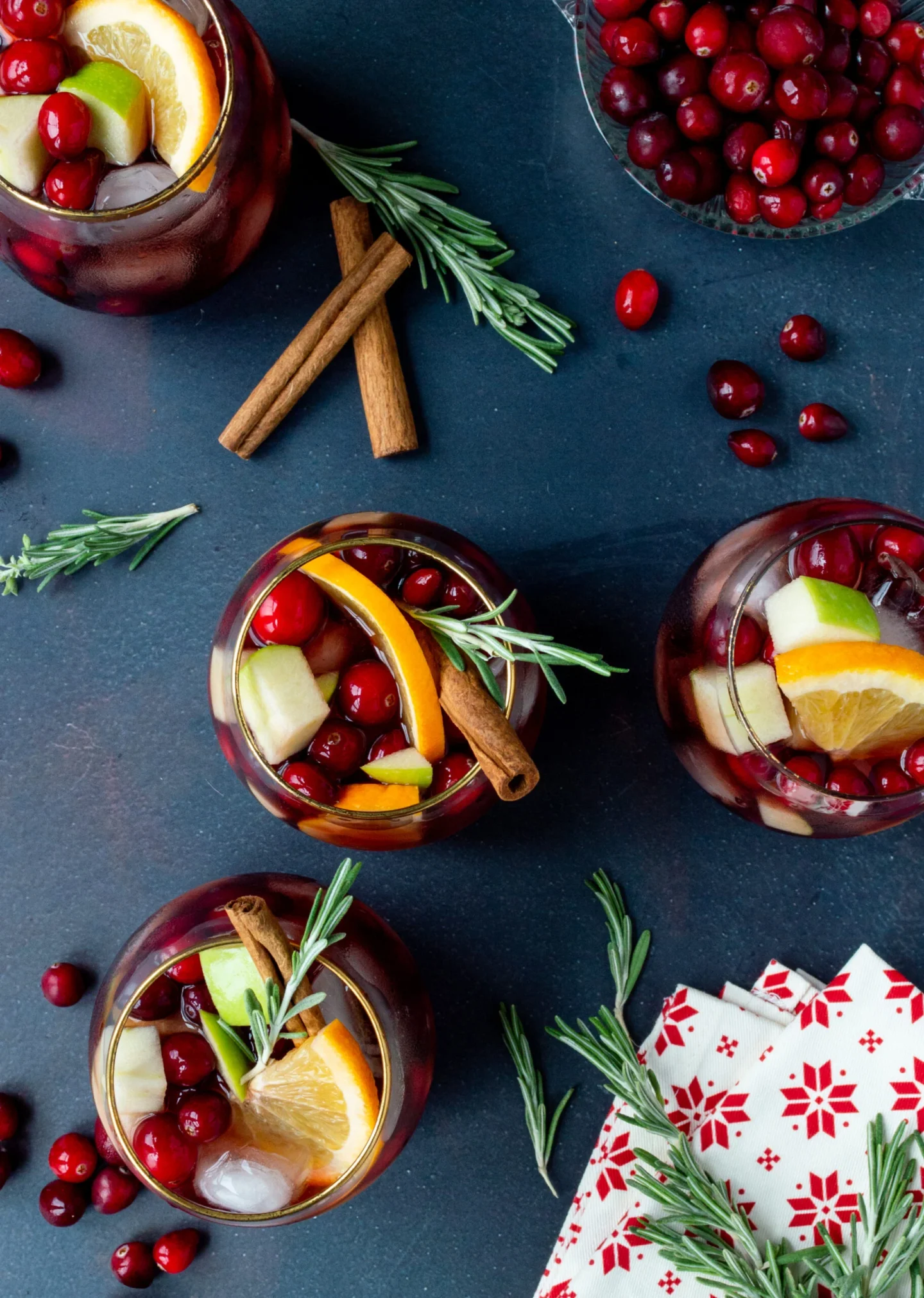 Four glasses of fruit-filled sangria garnished with orange slices, cranberries, rosemary, and cinnamon sticks rest on a dark surface alongside extra cranberries and festive holiday napkins—perfect for Autumn Gatherings or anyone searching for Cozy Fall Drinks.