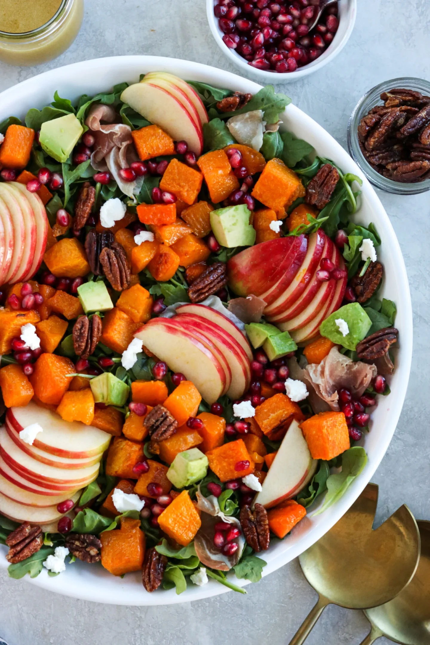 A bowl of fresh salad featuring cubed roasted squash, sliced apples, pomegranate seeds, pecans, avocado, and prosciutto, with side bowls of dressing, pomegranate seeds, and pecans.