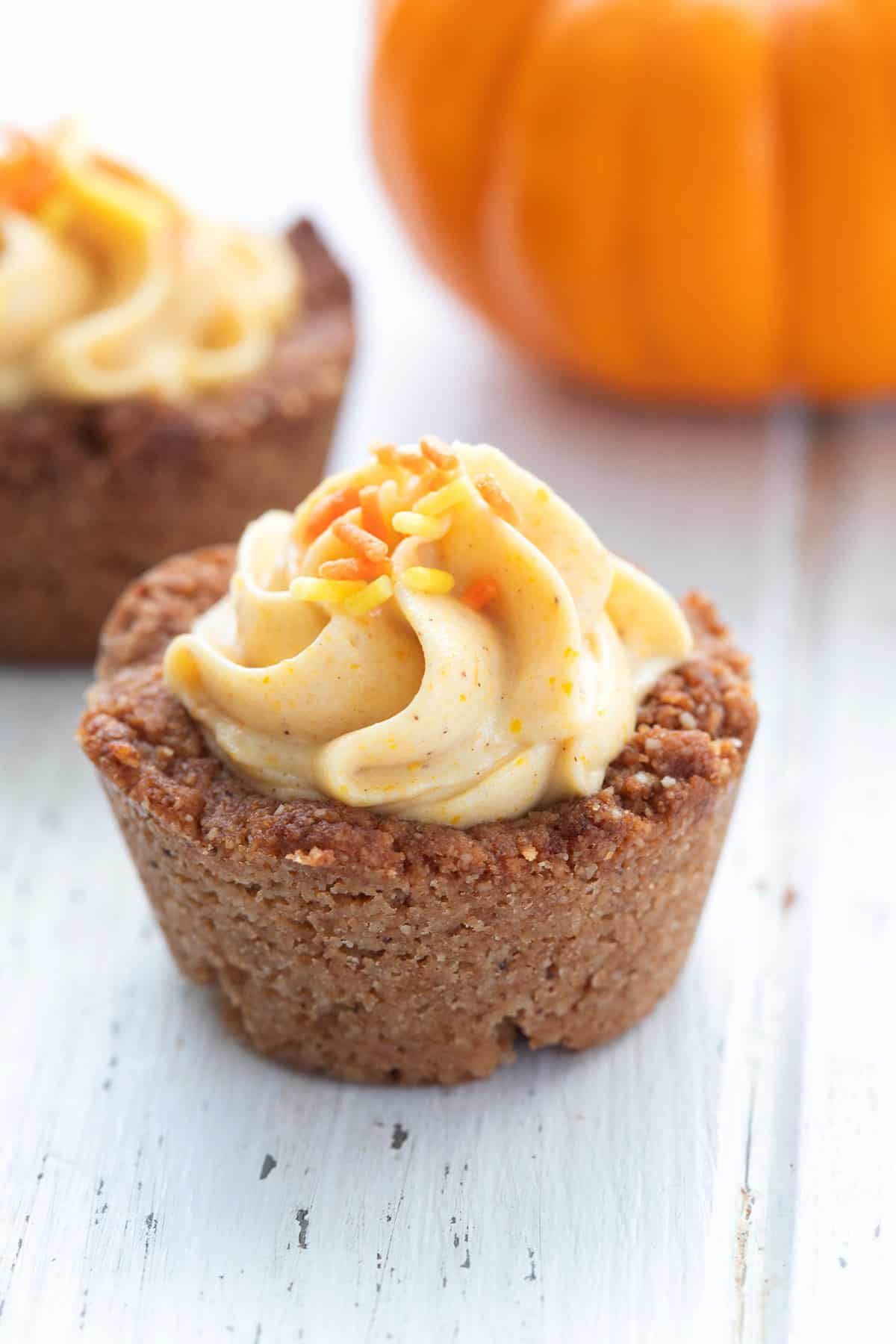 Two mini pumpkin tarts topped with cream frosting and sprinkles, serving as healthy Thanksgiving desserts with a whole pumpkin in the background.