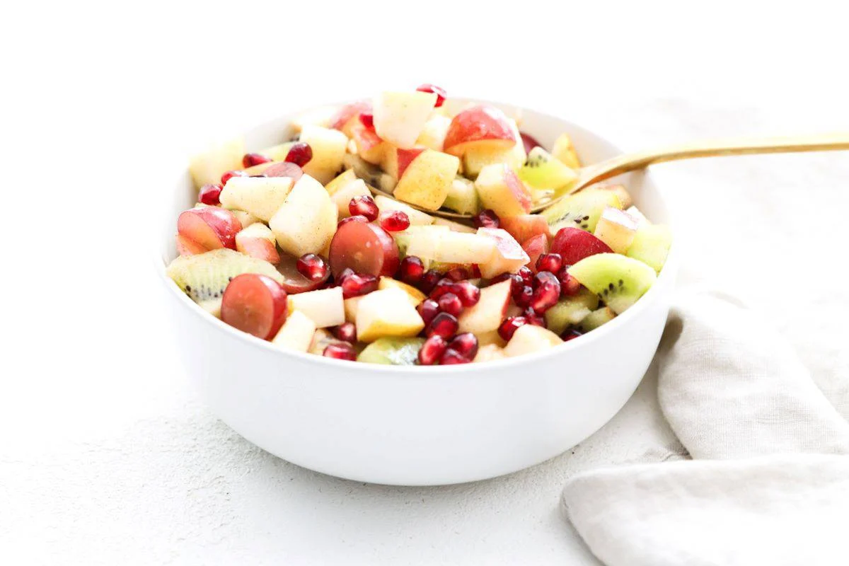 A white bowl filled with a healthy fruit salad of chopped apples, grapes, kiwi, and pomegranate seeds.
