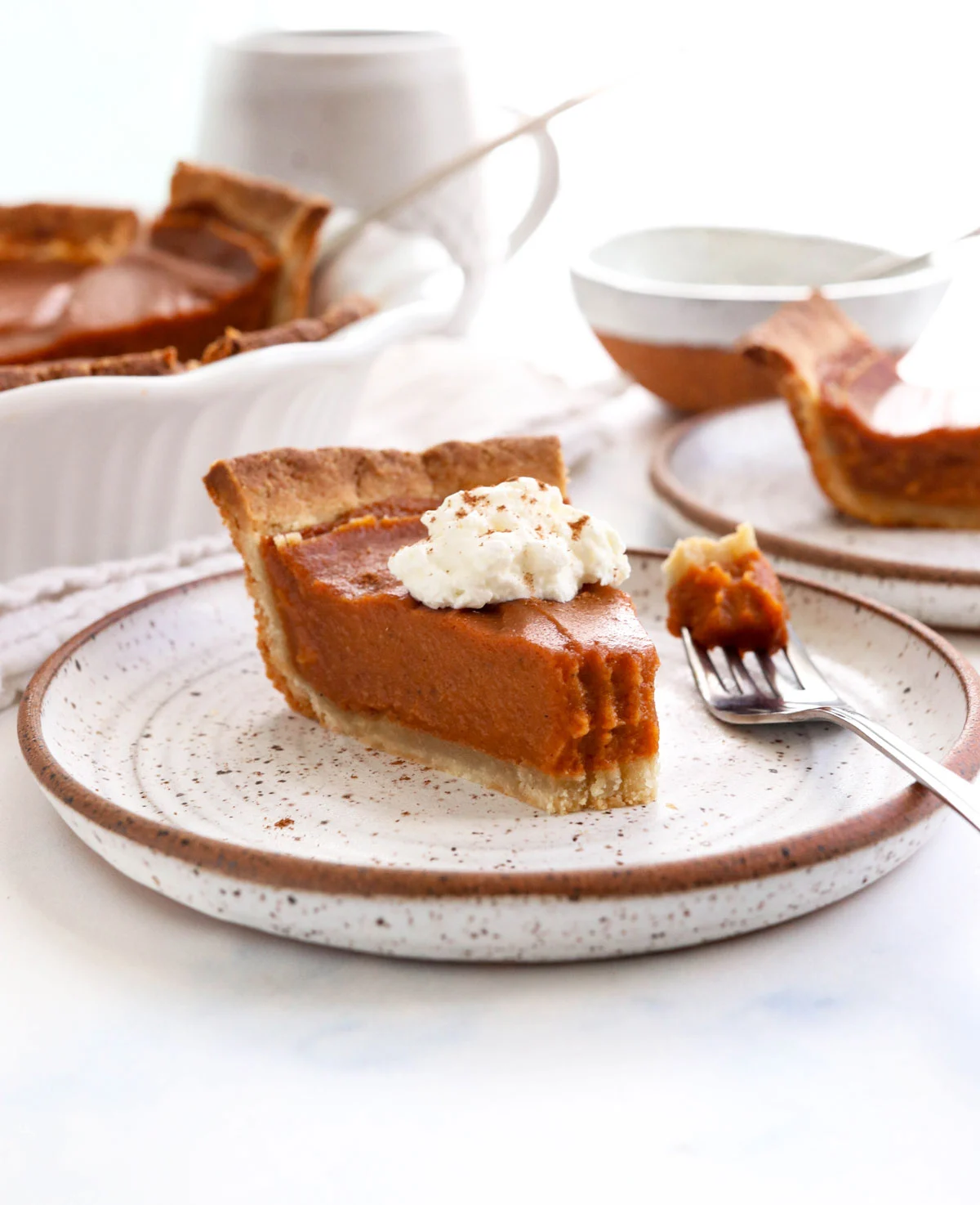 A slice of pumpkin pie topped with whipped cream on a speckled plate, with a fork and a whole pumpkin dessert in the background.