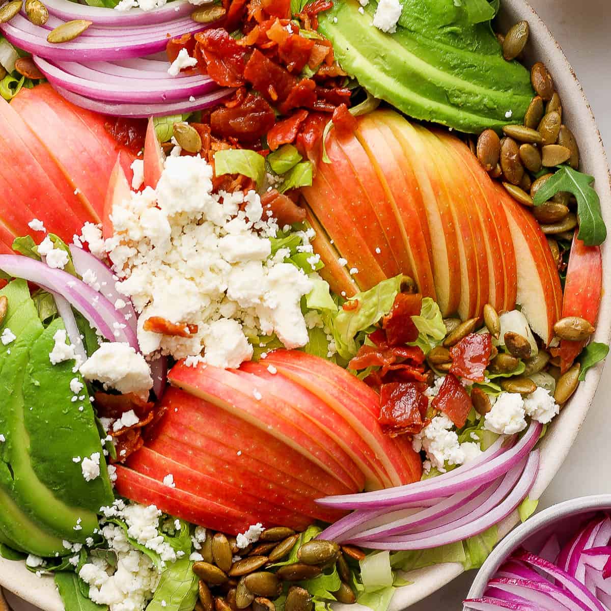 A bowl of fresh salad topped with sliced apples, avocado, red onion, crumbled feta cheese, bacon bits, and pumpkin seeds, perfect for healthy Thanksgiving recipes.