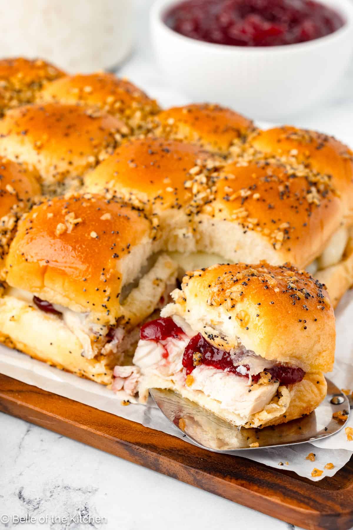 A spatula lifting a turkey and cranberry slider from a tray of Thanksgiving leftover recipes topped with poppy seeds.